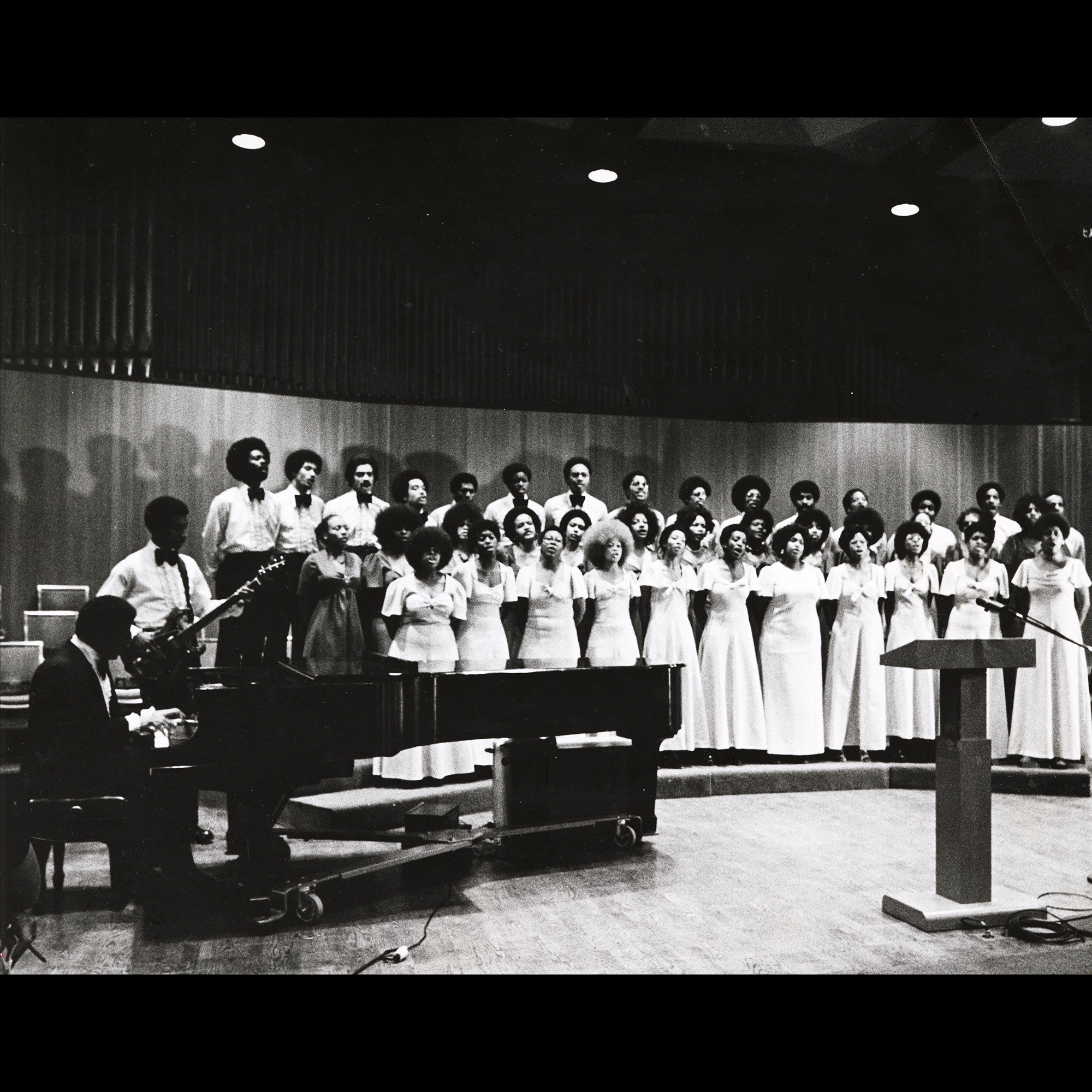 Black and white photo of an early Sounds of Blackness performance. Singers stand on risers with a pianist and guitarist to the side.