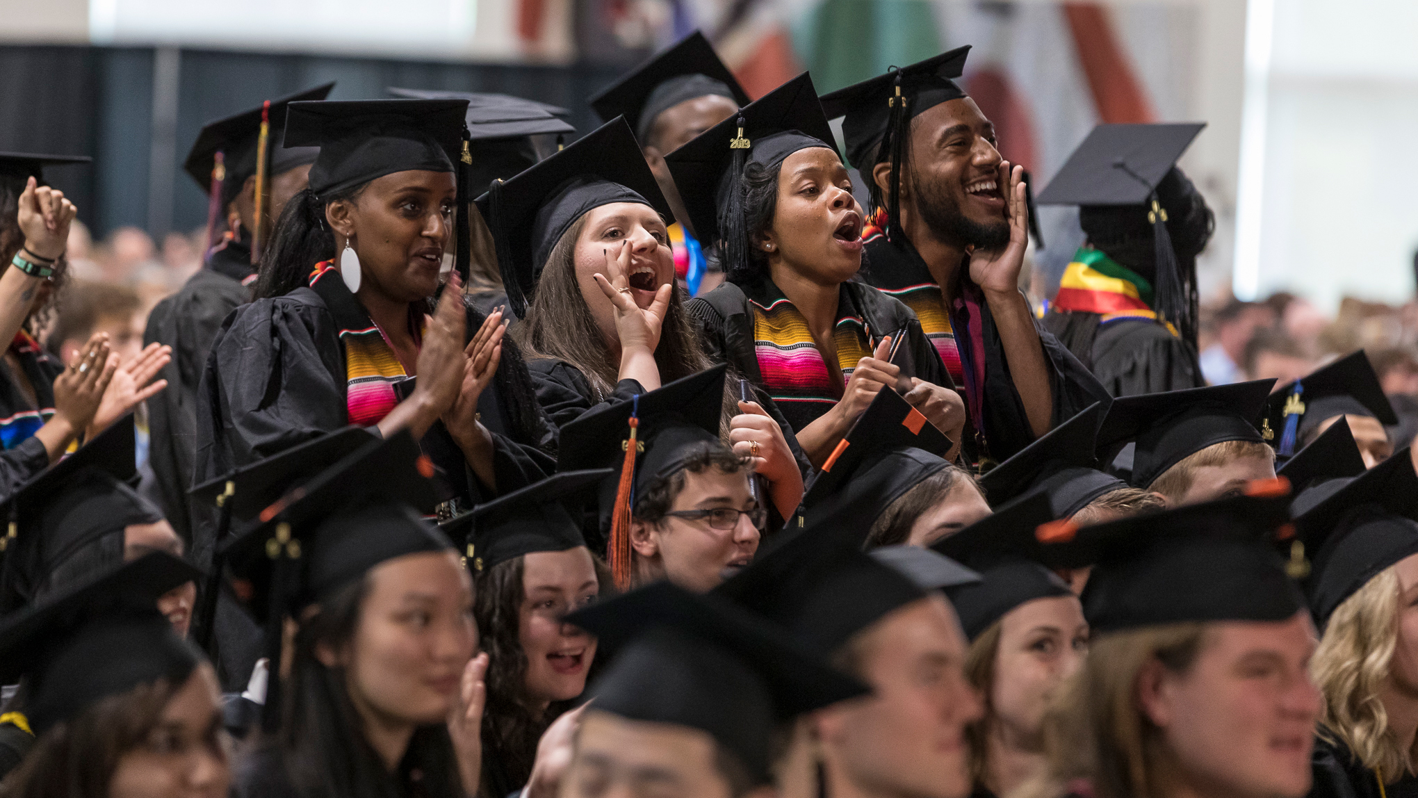 Photo of students cheering in graduation caps and gowns standing up and cheering