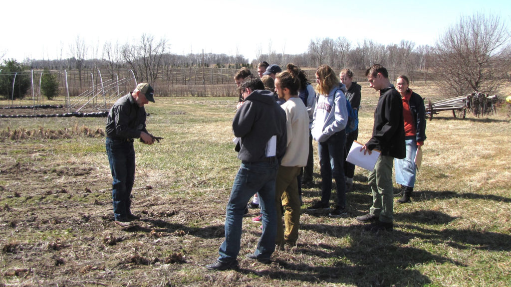 A group of students stand in a field watching a farmer speak