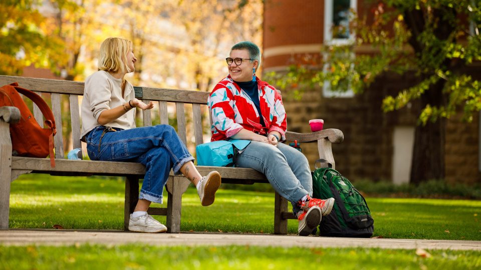 Photo of two students talking on a bench near the library building.