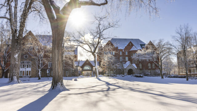 Macalester's Great Lawn covered in snow. The trees are bare, and Old Main, a historic brick building has snow on its roof.