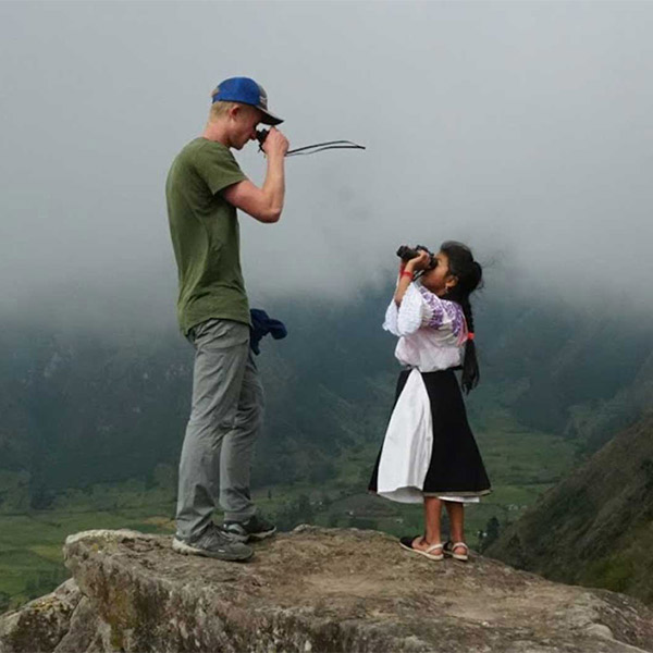 A student with a local inhabitant standing upon a mountain looking at each other with binoculars.