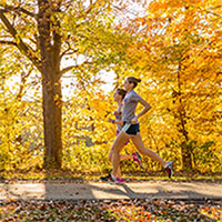 Photo of two students running in a park in front of trees with yellow leaves.
