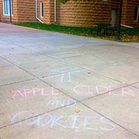 Photo of a sidewalk with chalk on it reading "APPLE CIDER AND COOKIES"