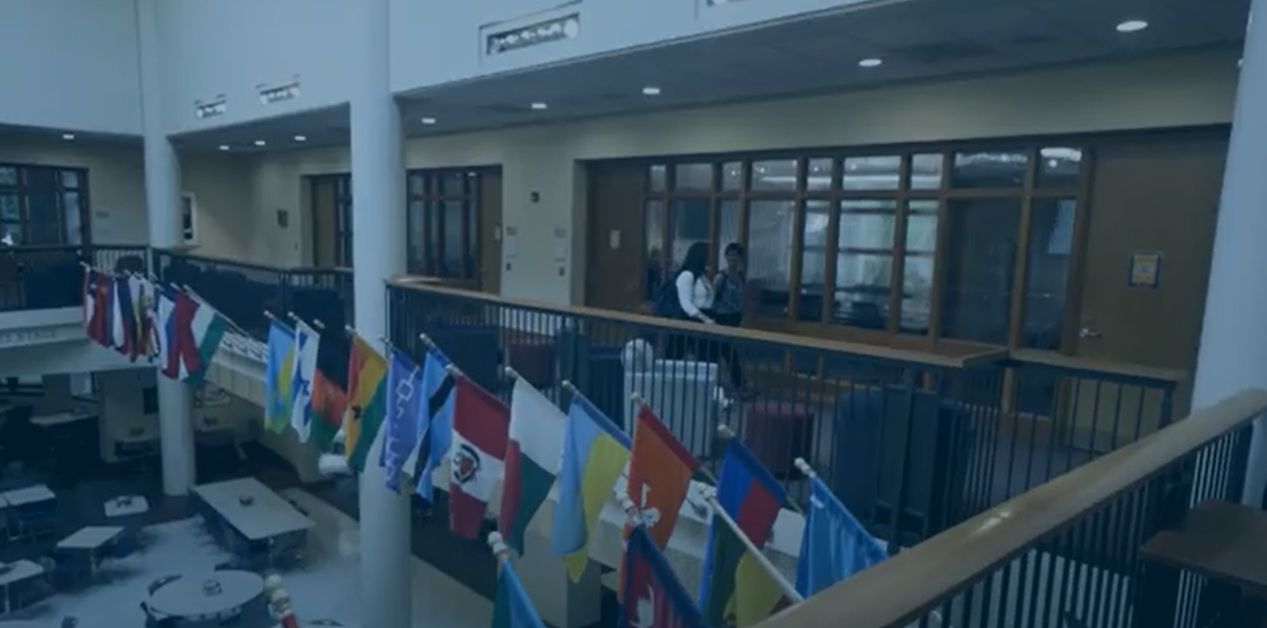 Two Macalester students walk across the top level of the Campus Center, passing the long row of flags from different countries that hang from the railing.
