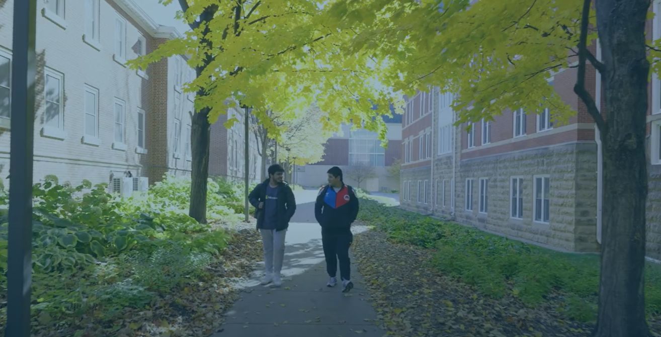 Two students walk through Macalester's campus, bordered on both sides by green foliage and red brick buildings.