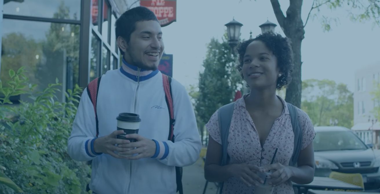 Two Macalester students walk toward the camera on Grand Avenue, holding coffee cups and chatting.