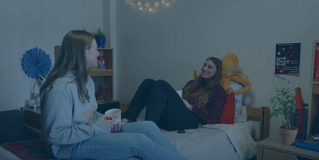 Two students sit on a dorm bed, chatting and smiling.
