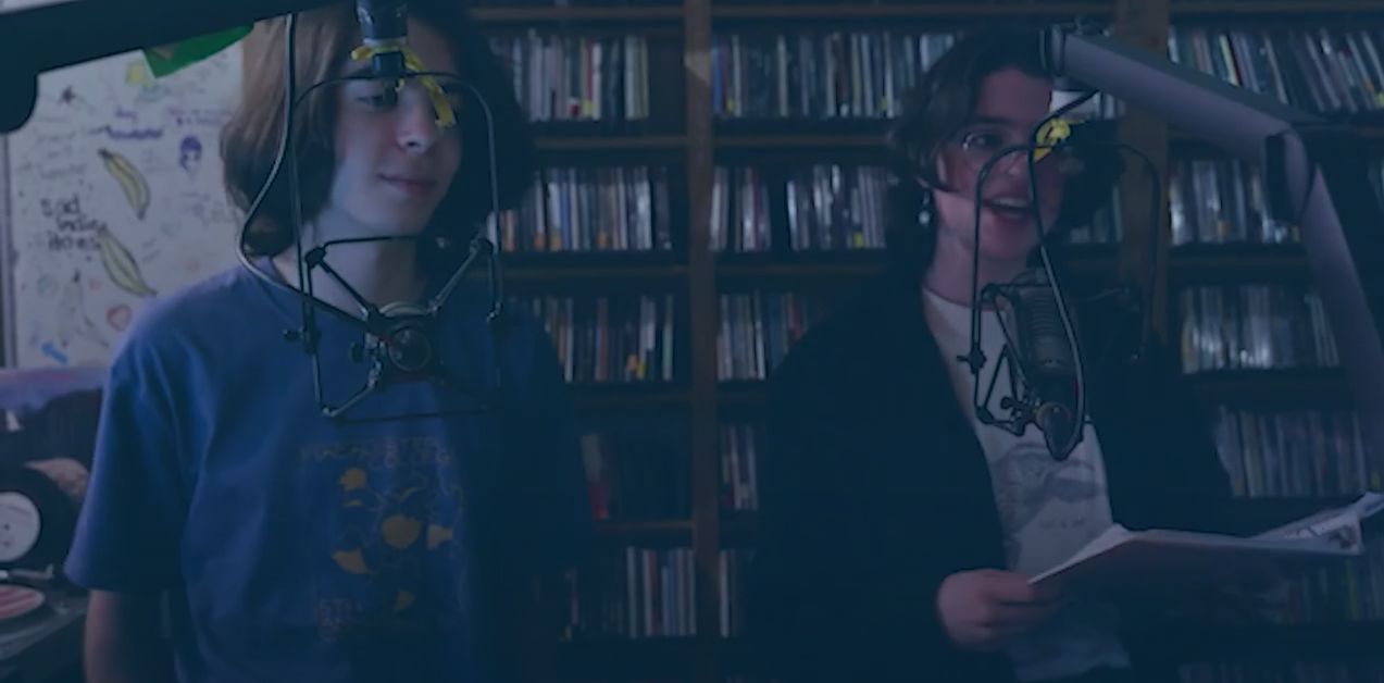 Two students speak into recording equipment, surrounded by CDs and vinyl records.