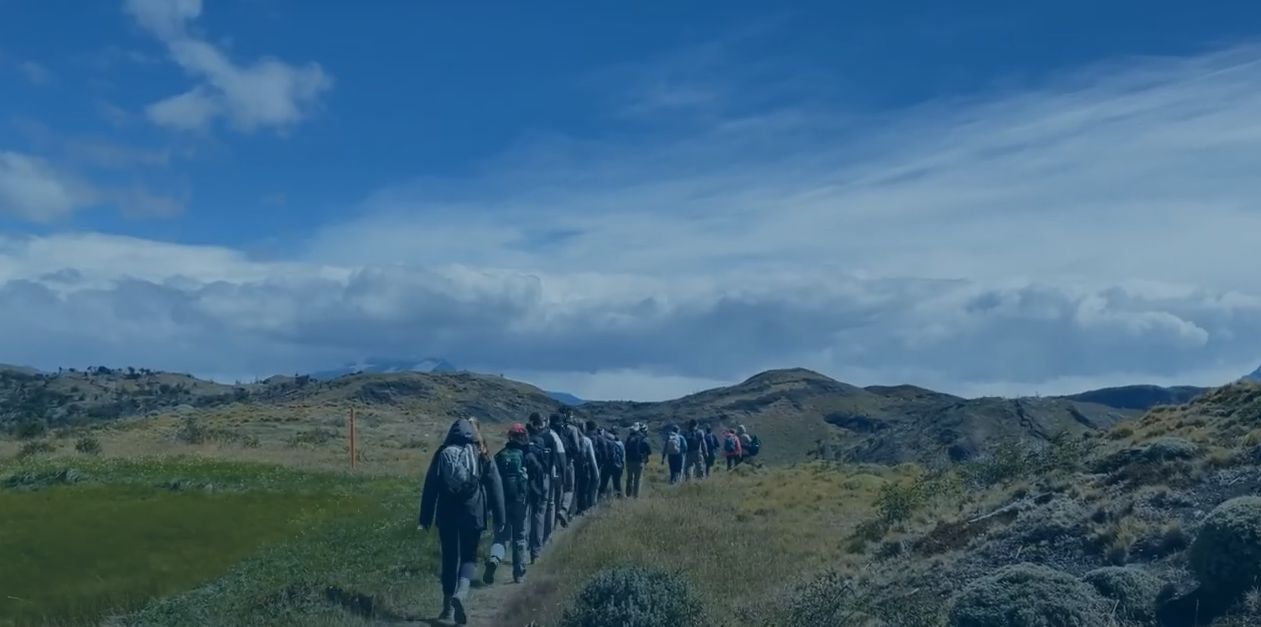 A long line of students walks across an open plain, wearing backpacks and outdoor gear. The blue sky above is clear and expansive.