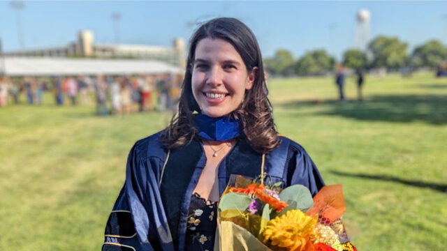 Ava Bindas '17 in a graduation gown, holding a bouquet of flowers outdoors on a sunny day.