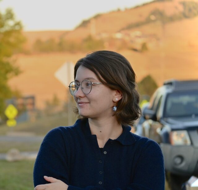 Macalester alum Chloe Moore '24 standing outdoors with arms crossed, with cars and hills in the background