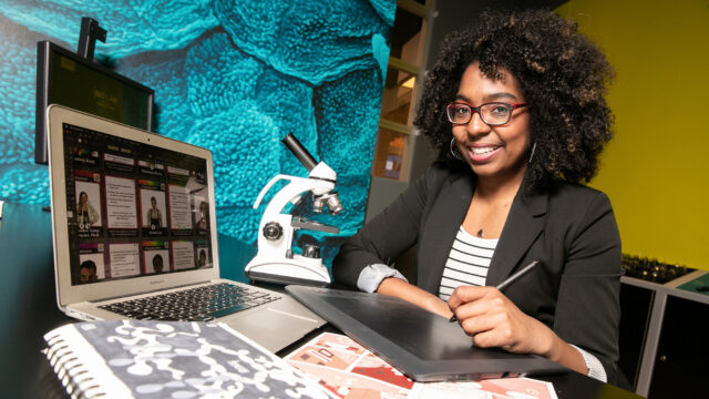 Jaye Gardiner ’11 sits at a desk with a laptop as she illustrates medical cartoons