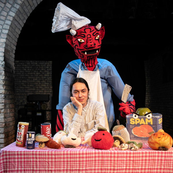 Jennings Mergenthal ’21 poses at a table covered with plush food props and canned goods, while a large red devil puppet dressed as a chef stands behind them holding a cleaver.