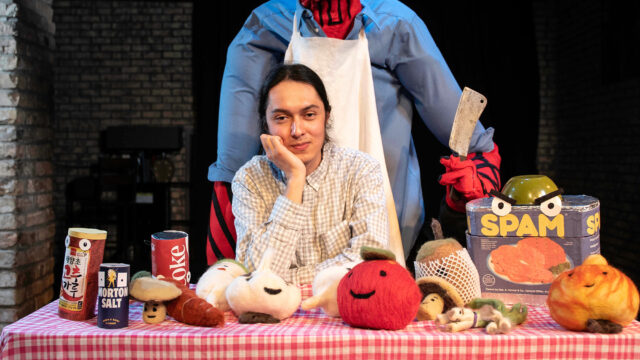 Jennings Mergenthal ’21 poses at a table covered with plush food props and canned goods, while a large red devil puppet dressed as a chef stands behind them holding a cleaver.