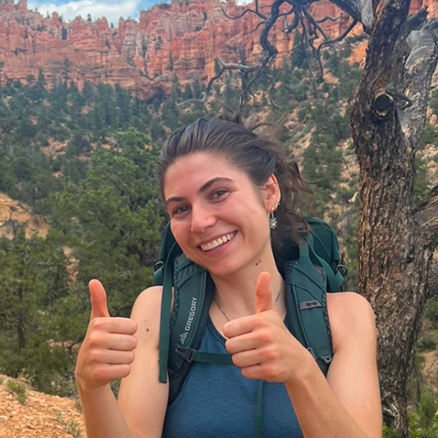 Julia Ricks ’22, wearing a backpack and giving two thumbs up, smiles on a hiking trail in front of a backdrop of red rock canyons and green trees