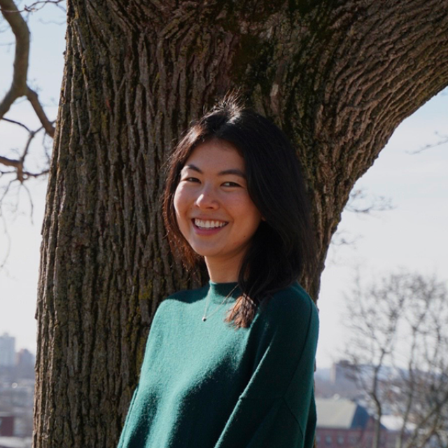 Karintha Lowe ’16 smiles while leaning against a large tree trunk on a sunny day. She is wearing a dark teal sweater, with blurred buildings visible in the background.