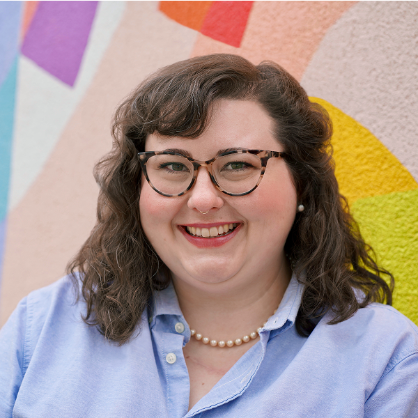 Kate D. Gallagher ’16 smiles, wearing a blue button-down shirt, pearl necklace, and tortoise-shell glasses, against a colorful mural background.
