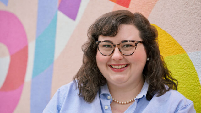 Kate D. Gallagher ’16 smiles, wearing a blue button-down shirt, pearl necklace, and tortoise-shell glasses, against a colorful mural background.