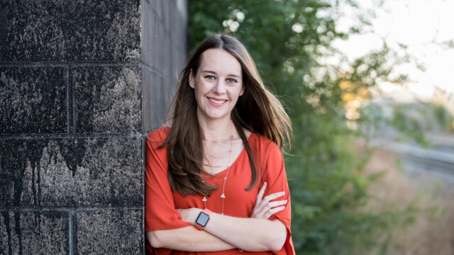 Kate Agnew ‘11, Senior Director of Engineering at Marriott International, leans against a dark concrete wall with her arms crossed, smiling brightly.
