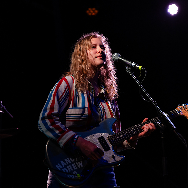 Kerry Alexander ’12, front person for Bad Bad Hats, is playing a blue electric guitar and singing into a microphone on a darkly lit stage.