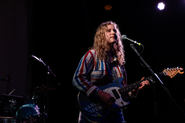 Kerry Alexander ‘12 plays guitar and sings. She is standing behind a microphone.