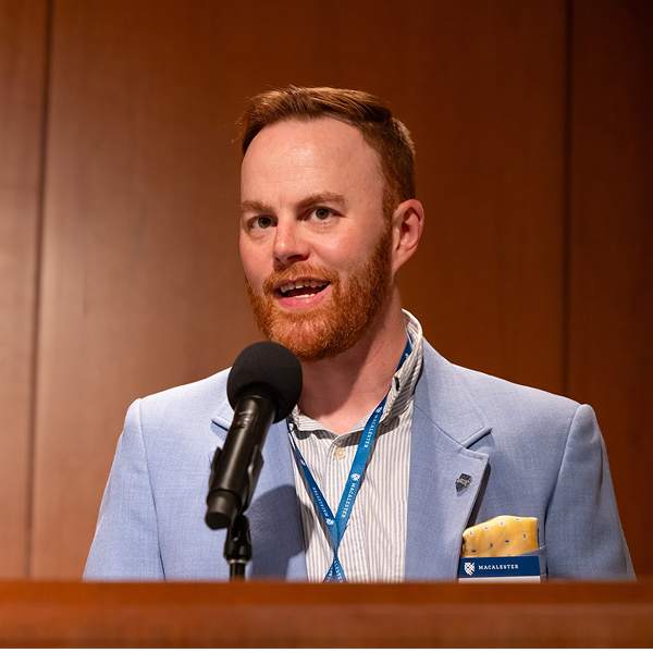 Kevin Finnegan ’11, wearing a light blue blazer, speaks into a microphone at a podium with a wood-paneled background.