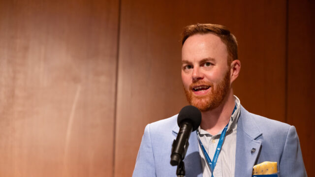 Kevin Finnegan ’11, wearing a light blue blazer, speaks into a microphone at a podium with a wood-paneled background.