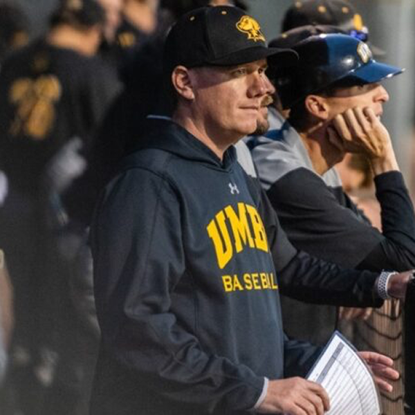 Liam Bowen ’06, Head Baseball Coach at UMBC, stands in the dugout wearing a black and yellow team sweatshirt and baseball cap, intently watching the game.