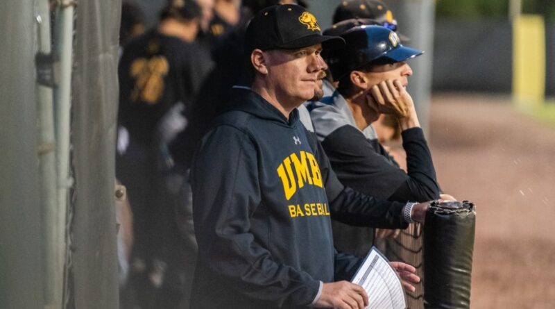 Liam Bowen ’06, Head Baseball Coach at UMBC, stands in the dugout wearing a black and yellow team sweatshirt and baseball cap, intently watching the game.