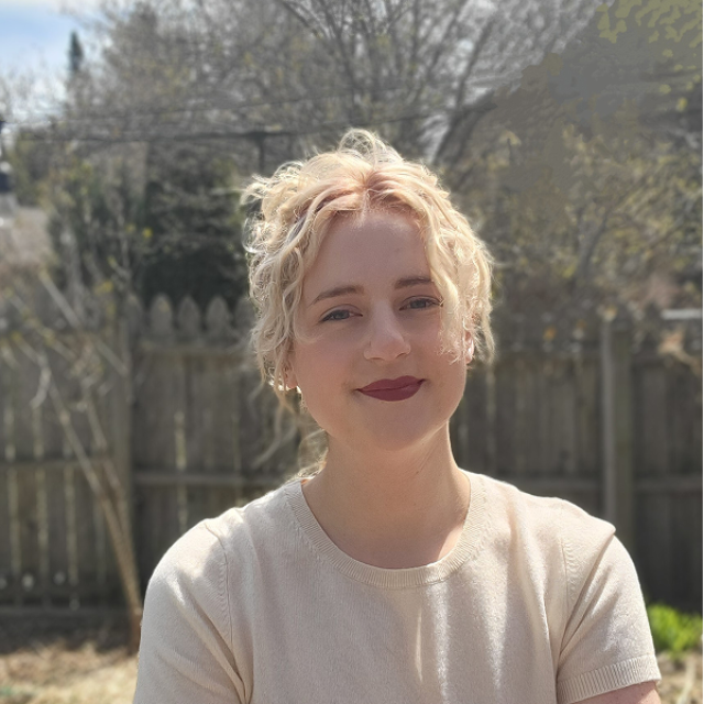 Madeline Stone Kutis ’14, with short, curly blonde hair, smiles in a headshot taken outdoors against a blurred wooden fence and trees.