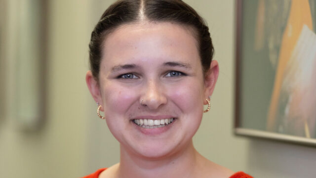 Mara Steinitz ’18, wearing a bright orange mesh-knit sweater, smiles in an indoor headshot