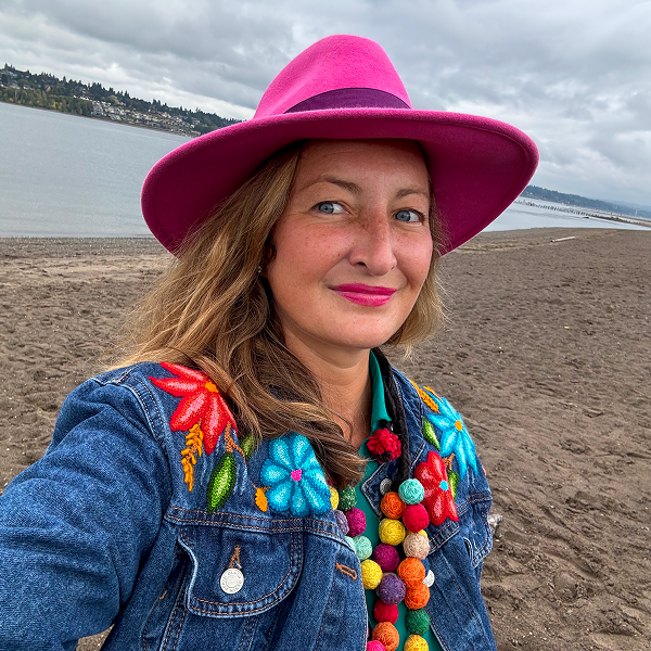 Marie Deschamps ’04, wearing a bright pink fedora and a denim jacket embroidered with vibrant flowers, smiles outdoors on a sandy beach.