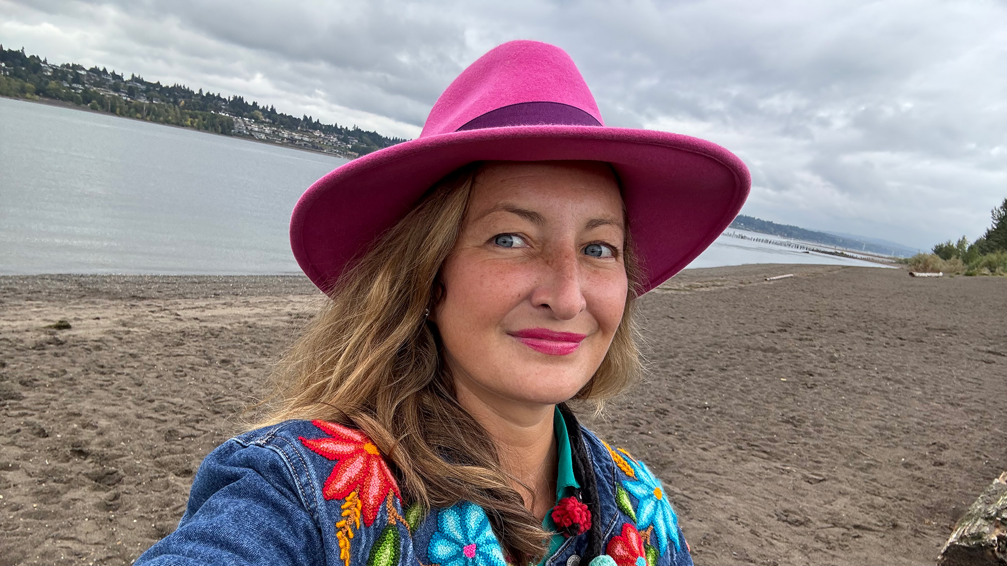 Marie Deschamps ’04, wearing a bright pink fedora and a denim jacket embroidered with vibrant flowers, smiles outdoors on a sandy beach.
