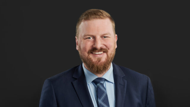 Matthew Bergeron ’08, a Shareholder at Larkin Hoffman, smiles in a professional headshot wearing a navy suit and a blue patterned tie against a dark background.