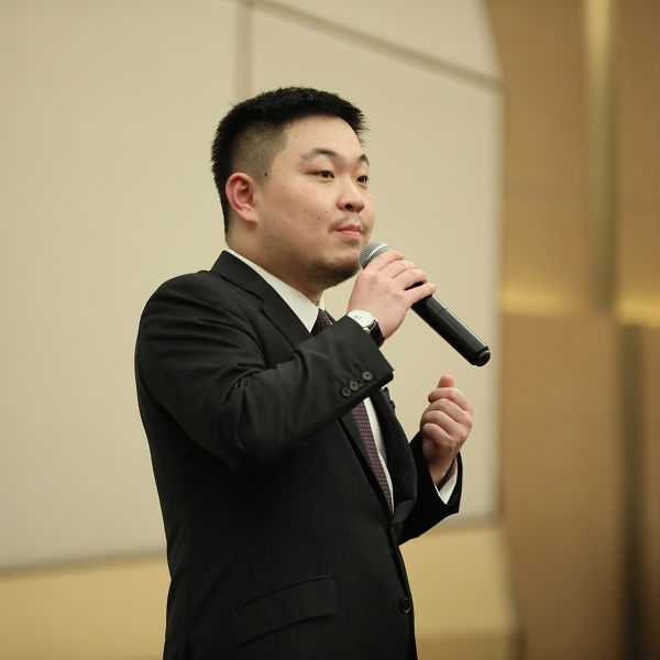 Max Wang ’15, Partner at Stooges Education Group, stands at a formal event, speaking into a microphone while wearing a dark suit.