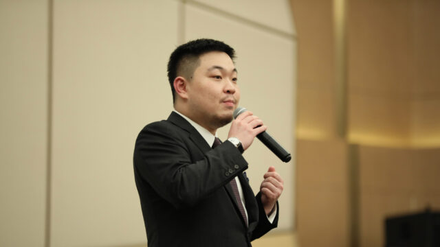 Max Wang ’15, Partner at Stooges Education Group, stands at a formal event, speaking into a microphone while wearing a dark suit.