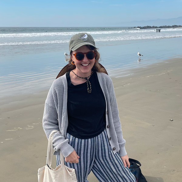 Megan Ritchie ’09, a television writer, smiles on a sunny beach, wearing sunglasses, a baseball cap, and a gray cardigan over a striped pants.