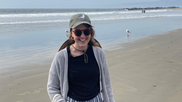 Megan Ritchie ’09, a television writer, smiles on a sunny beach, wearing sunglasses, a baseball cap, and a gray cardigan over a striped pants.