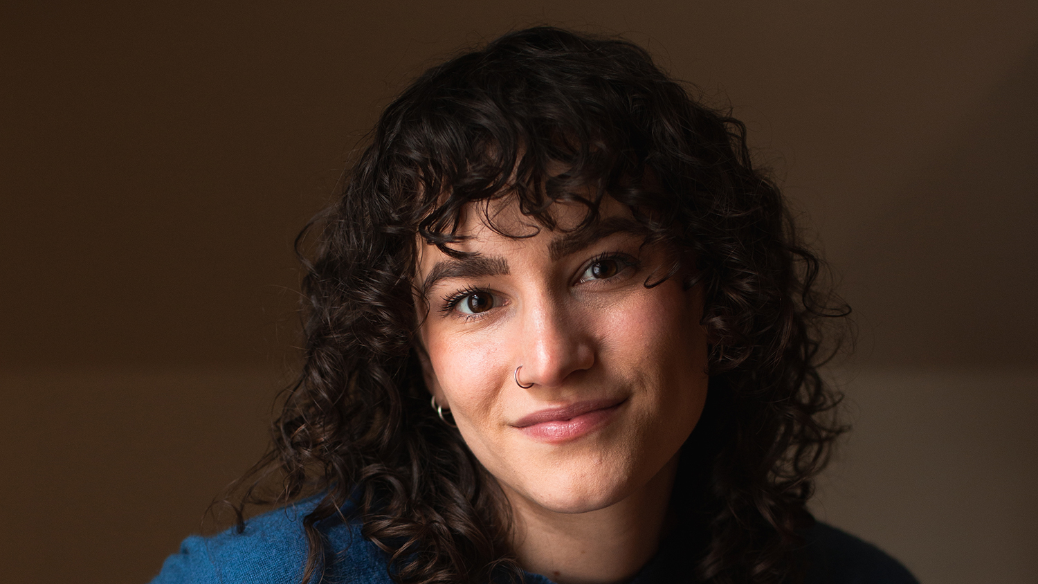 Miriam Moore-Keish ’19, Content Strategist at Capstone Publishing, smiles in a headshot with curly dark hair and a nose ring, wearing a blue sweater.