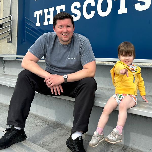 Tony Armstrong ’13 smiles while sitting on bleachers with a young child pointing toward the camera, below a sign reading 'THE SCOTS'.