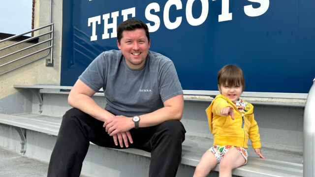 Tony Armstrong ’13 smiles while sitting on bleachers with a young child pointing toward the camera, below a sign reading 'THE SCOTS'.