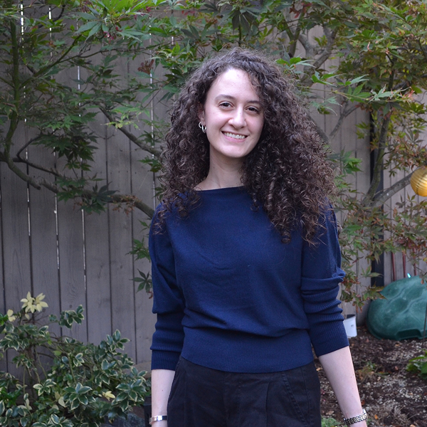 Zeena Fuleihan ’18, a PhD candidate, smiles outdoors with long, dark curly hair, wearing a navy sweater against a wooden fence and greenery.
