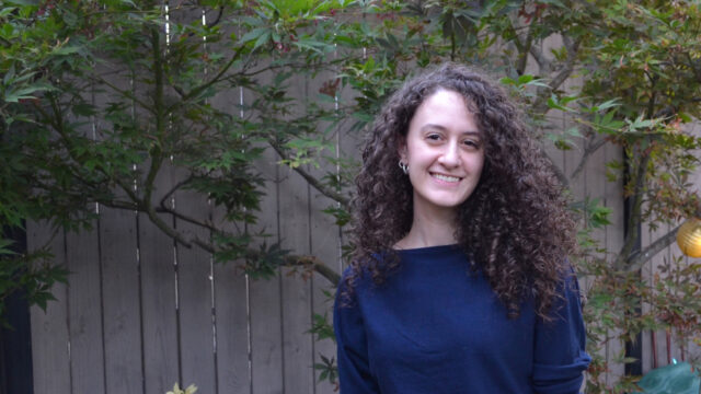 Zeena Fuleihan ’18, a PhD candidate, smiles outdoors with long, dark curly hair, wearing a navy sweater against a wooden fence and greenery.