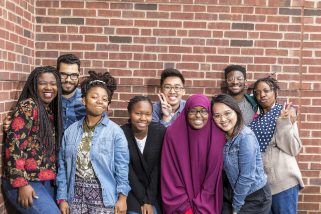 A group of nine students stands in front of a brick wall smiling for a picture