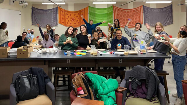 Twenty students sit at and stand behind a table, smiling and posing for a picture while holding decorated sugar cookies.