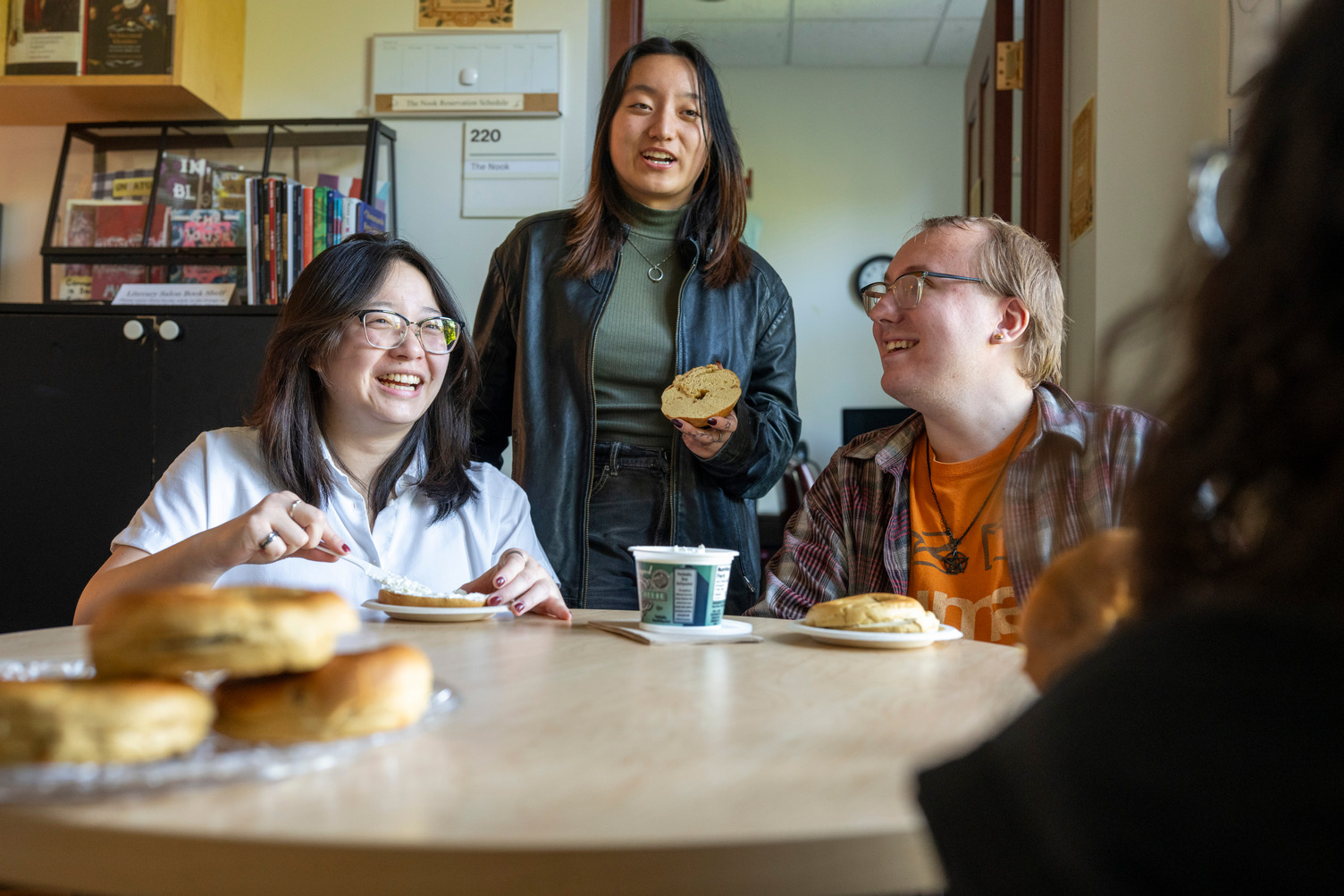 Two students sit at a small table in the English department office space, while one stands behind them. All three are laughing and eating bagels, or have a bagel on their plate. Another student sits with their back to the camera, just out of focus.