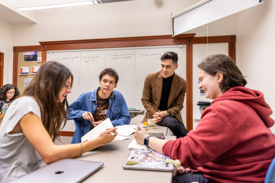 Three students sit at a table in English class, animatedly discussing something while surrounded by laptops and notepads. A professor sits on the edge of the table, listening to the conversation.