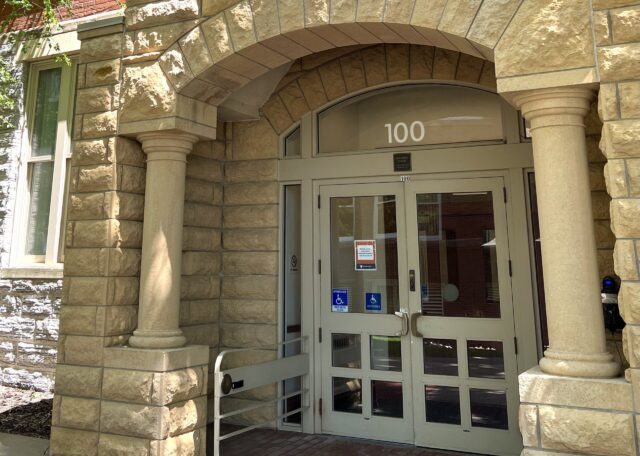 The west entrance to the Old Main building. The tan brick façade creates an arch over the double glass door entrance. There is a small black card reader on a handrail a couple feet extended from the left door handle, next to the ADA accessible door opener button.