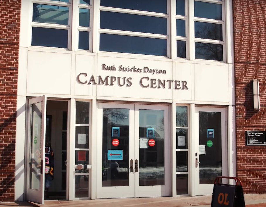 The exterior of the building with its name Ruth Striker Dayton Campus Center above a set of double doors with a single door on either side. The left single door is ajar as someone enters.